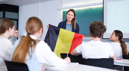 Teenage students sitting in class and listening carefully to female teacher holding Belgian flag in hands