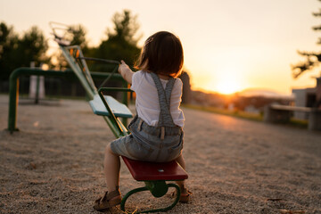 back view of one small caucasian toddler child sitting alone on the seesaw in park in sunset lonely...