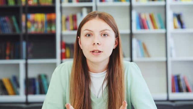 Teenager girl with long hair talking emotionally to webcam and gesturing during online meeting, blurred bookshelve background. Closeup of young female expressing opinion. Concept of communication