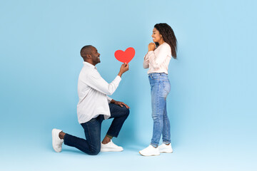 Romantic african american man on his knees giving red heart shaped card to excited black woman, side view
