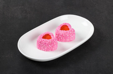 Vegan festive dessert. Small Strawberry cream mousse cake with jelly filling, in the shape of a heart, sprinkled with pink coconut chips. On a plate. Dark gray background. Valentine's day