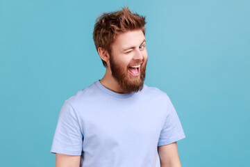 Portrait of bearded man being in good mood, smiling broadly and winking at camera, keeps mouth...