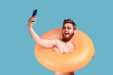 Portrait of excited bearded man posing with orange rubber ring and holding mobile phone, making selfie, enjoying his vacation at seaside. Indoor studio shot isolated on blue background.