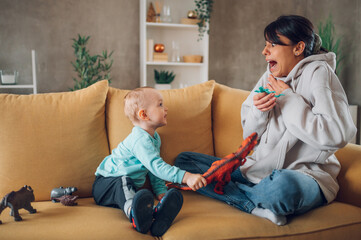 Mother and toddler son playing with dinosaurs toys at home on the couch