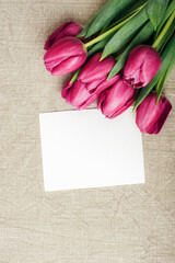 Purple tulips and blank card on gray tablecloth. Spring holidays concept. Top view, flat lay, mockup