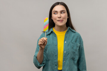 Positive woman with dark hair holding multicolored ice cream in hands, feels hungry, wants to taste dessert, wearing casual style jacket. Indoor studio shot isolated on gray background.