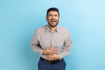 Portrait of funny handsome bearded young businessman standing with closed eyes and laughing, holding his belly, wearing striped shirt. Indoor studio shot isolated on blue background.