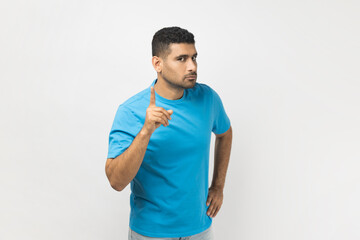 Portrait of serious bossy attractive unshaven man wearing blue T- shirt standing with raised index finger, warning somebody, showing disapproval gesture. Indoor studio shot isolated on gray background