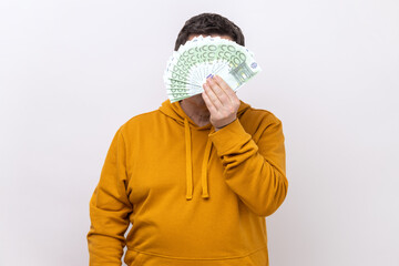 Unknown man hiding face behind fan of hundred euro bills, big winning, betting, wearing urban style hoodie. Indoor studio shot isolated on white background.