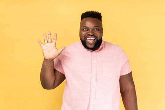 Portrait of friendly bearded man in pink shirt greeting you rising hand and waving, saying hi, glad to see you, looking at camera with toothy smile. Indoor studio shot isolated on yellow background.