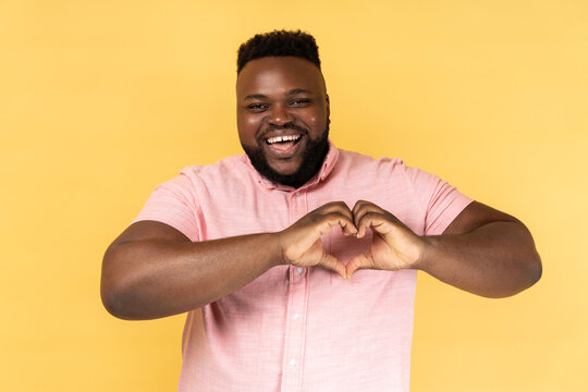 Portrait of lovely romantic happy man wearing pink shirt making heart shape with fingers, gesturing love hope charity sign, looks at camera. Indoor studio shot isolated on yellow background.