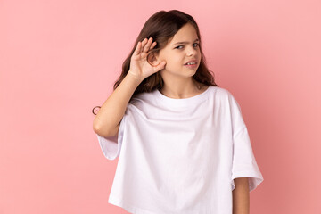 Portrait of curious little girl wearing white T-shirt holding hand near ear trying to hear secrets and find out gossips, listen to private whispers. Indoor studio shot isolated on pink background.