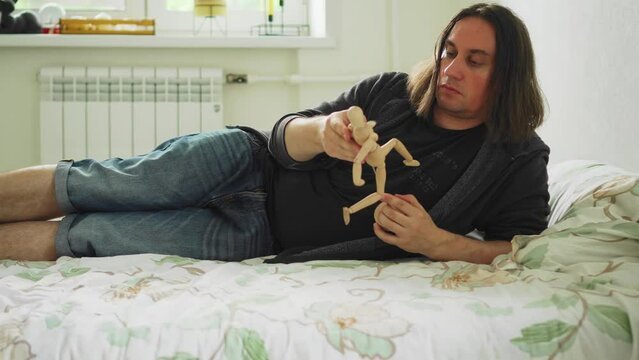 The guy is lying on the bed with a wooden figure of a man in his hands. He moves his limbs