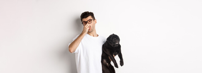 Young man shut nose as holding pug, disgusted with bad smell fart of animal, standing over white background