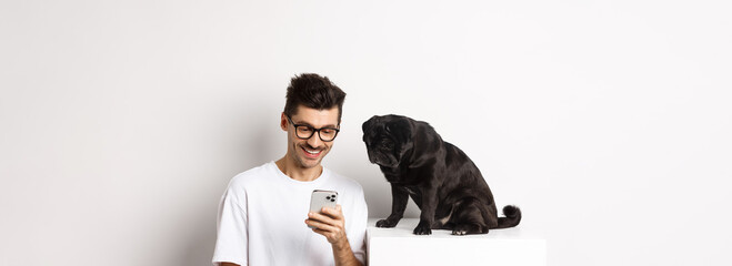 Smiling young man using smartphone and sitting near dog. Pug owner checking photos on mobile phone,...