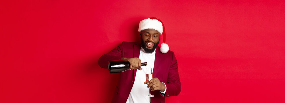 Christmas, Party And Holidays Concept. Handsome Black Man In Santa Hat Pouring Glass Of Champagne And Smiling, Celebrating New Year, Red Background