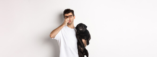 Young man shut nose as holding pug, disgusted with bad smell fart of animal, standing over white...