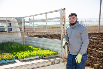 Portrait of confident young bearded farmer standing near truck with vegetable seedlings on farm field on sunny spring day