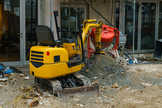 Small yellow excavator and concrete mixer at a construction site in front of a building under construction.