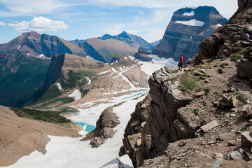 GLACIER NATIONAL PARK, MONTANA, USA. A man in a red t-shirt stands atop a cliff overlooking a glacier and alpine scene of rocks, snow, and peaks.
