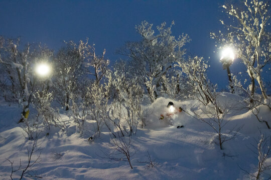 A male freerider is skiing in twilight on a slope in the ski resort Niseko United. Next to the deep powder snow, Hokkaido is known for great night skiing. His head lamp shines thro