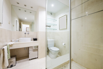 Beautiful bathroom with white marble tiles with a mirror integrated into the wall and porcelain toilets, wall lights in the false ceiling, glass shelves and a cabinet with a mirror under the sink