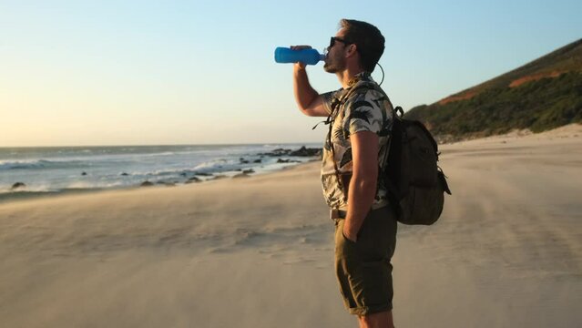 A traveler takes pictures with a modern camera of landscapes on the ocean shore among rocks and waves. male traveler with backpack drinking water on ocean beach at sunset. sport and travel vacation