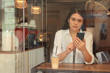 Young beautiful woman with smartphone in cafe, view through window glass