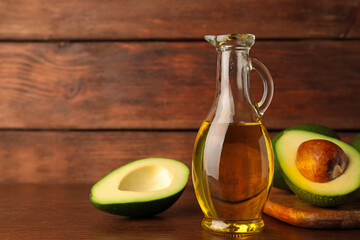 Glass jug of cooking oil and fresh avocados on wooden table, space for text