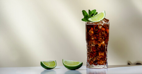 Whiskey cola cocktail with strong alcohol and ice, garnished with mint and lime in highball glass. Beige background, hard light - obrazy, fototapety, plakaty