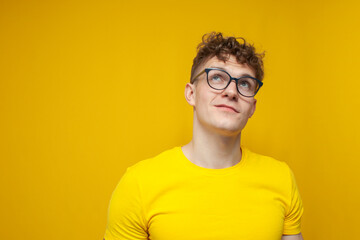curly pensive guy in glasses in a yellow t-shirt is dreaming and looking up on a yellow background
