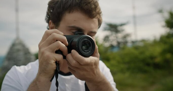 Close up of a young man holding a camera, taking a picture and than taking a look without the camera. Green background with the trees and cloudy sky.