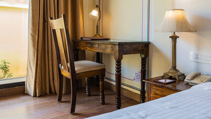 The workplace in the room in a retro style. Lamps with shades stand on a wooden table and a bedside table. Paper, pencil, folder on the countertop. A chair with a padded seat. Curtains on the window.