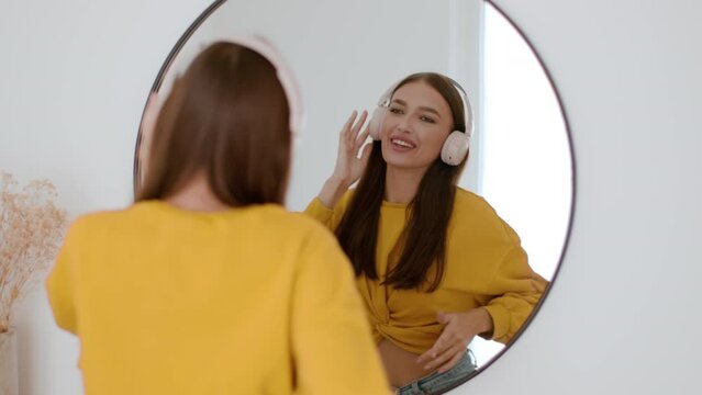 Young happy carefree woman wearing wireless headphones enjoying favorite music and dancing, looking at mirror at home