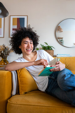 Happy African American woman relaxing at home reading a book sitting on the couch looking at camera. Vertical