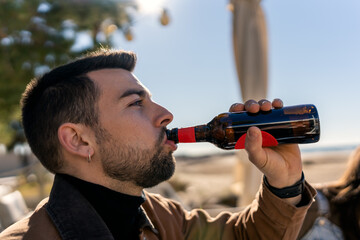 Side view of young bearded guy drinking beer from glass bottle in sunlight while spending weekend on beach