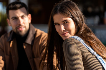 Positive young female in casual clothes with long dark hair looking at camera while sitting with male friend in outerwear on street