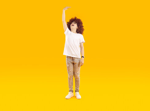 Little boy wishes he were taller. Happy kid raises his hand to show how tall he wants to grow. Full body child in casual T shirt, jeans and funny wig measuring his height isolated on orange background