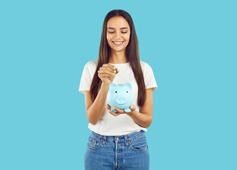 Portrait of happy woman who puts coin in piggy bank, saving money for purchase she dreams of. Young...