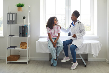 Caring, understanding doctor touches shoulder of excited teenage girl who is worried before medical examination. African American girl is sitting on examination couch and talking to female doctor. - obrazy, fototapety, plakaty