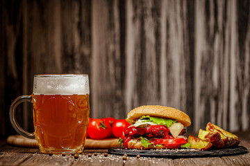 Front view of homemade beef burger with mug of beer, potato, tomatoes, cheese, on wood background.