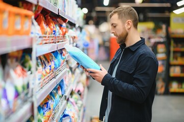 Young cheerful positive male customer making purchases in supermarket, buying household chemicals