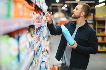 Young cheerful positive male customer making purchases in supermarket, buying household chemicals
