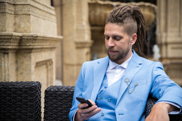 Young successful man in a blue suit looking at a smartphone while sitting in a luxury street cafe.
