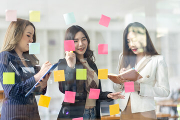 Young creative business Asian woman standing with her colleagues writing new ideas on sticky notes over glass wall, business marketing strategy or User experience ux ui concept.