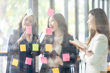 Young creative business Asian woman standing with her colleagues writing new ideas on sticky notes over glass wall, business marketing strategy or User experience ux ui concept.
