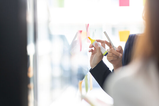 Young creative business Asian woman standing with her colleagues writing new ideas on sticky notes over glass wall, business marketing strategy or User experience ux ui concept.