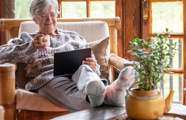 Smiling relaxed pensioner woman sitting at home in an armchair using a digital tablet to see videos...