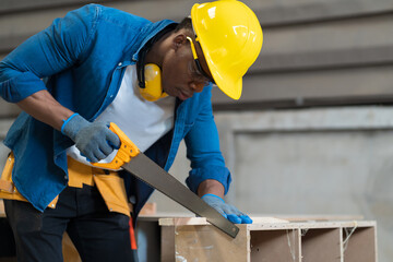 Professional African American male carpenter cutting lumber wood with handsaw in the wood workshop. Male carpenter working at the furniture workshop - Powered by Adobe