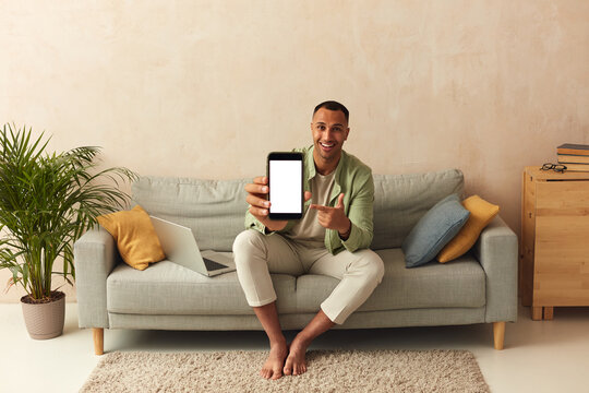 Man Showing Smartphone At Home. Handsome Multiracial Guy Sitting On Sofa And Demonstrating Mobile With Blank White Screen In Cozy Living Room at Home